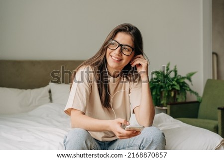 Young happy smiling woman sitting at home listening to music on phone headphones Royalty-Free Stock Photo #2168870857