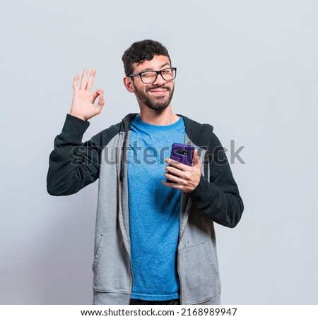 Pleasant person holding cellphone and winking, smiling boy with cellphone making ok sign, concept of positive person with cellphone closing fingers in ok sign Royalty-Free Stock Photo #2168989947