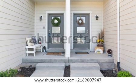 Panorama Two gray front door of a townhouse with two concrete entrance way. Home entrance with decorated doors with wreath and glass panels in the middle of a wall with vinyl wood and stone Royalty-Free Stock Photo #2169007947