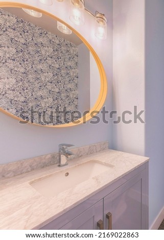 Vertical Powder blue bathroom interior with floral wall paper. There is a toilet with portrait picture frame at the back and a vanity sink with marble counter and framed oval mirror.