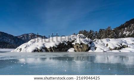 Picturesque boulders on the bank of a frozen river. Layers of snow on the rocks. Reflection on smooth turquoise ice. Mountains against the blue sky. Altai. Katun Royalty-Free Stock Photo #2169046425