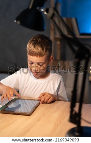 Boy looking at digital tablet while sitting at table at night, child reading e-book or watching cartoons using tablet, internet addiction