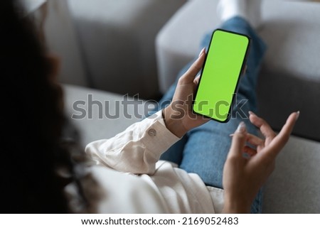 Young woman user customer hold smartphone mock up green screen in hand use mobile shopping app, check social media news, texting mobile sms order food delivery sit on sofa at home. Over shoulder view