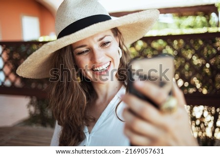 smiling modern housewife in white shirt with hat having video chat on a smartphone in the patio of guest house hotel.