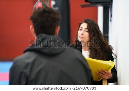 Young woman with yellow folder listening to sportsman while sitting on bench during motivation therapy session in modern gym Royalty-Free Stock Photo #2169070983