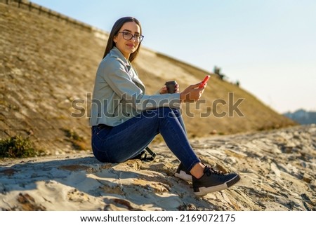Happy beautiful young woman sitting by the river while using a phone and holding coffee cup.