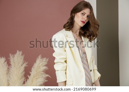 High fashion photo of a beautiful elegant young woman in cream white jacket, blazer, long dress, necklace, bracelets. Studio Shot. Rosy brown and Striped Corrugated Wall, dry branches.  