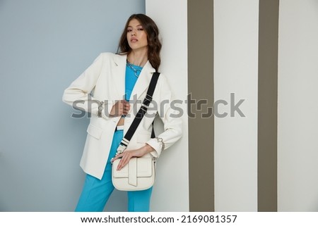 High fashion photo of a beautiful elegant young asian woman in a pretty white jacket, blazer, aqua blue top, pants, trousers, accessories, handbag. Striped Corrugated Wall. Studio Shot. 