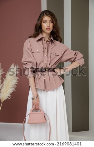 High fashion photo of a beautiful elegant young asian woman in pretty rosy brown shirt, belt, necklace, bracelets, white cream long skirt, handbag. Green striped Corrugated Wall, dry branches.