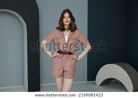 High fashion photo of a beautiful elegant young woman in pretty rosy brown jumpsuit, belt, necklace, bracelets. Bicolor wall, light and dark blue. Studio shot