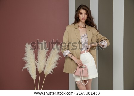High fashion photo of a beautiful elegant young asian woman in sandy brown jacket, blazer, cream skirt, top, necklace, bracelets. Studio Shot. Rosy brown and Striped Corrugated Wall, dry branches.   Royalty-Free Stock Photo #2169081433