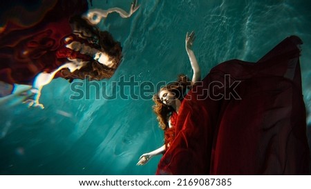 Photo underwater, a young beautiful woman in red with red hair reaches for the surface of the water, a human and his reflection. mystical underwater portrait