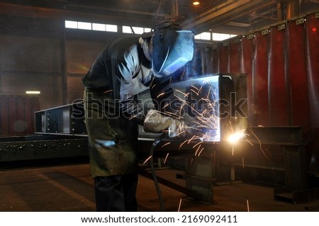 A welder in protective overalls and a welding mask welds metal structures by welding on a semi-automatic line, blue sparks fly to the sides Royalty-Free Stock Photo #2169092411
