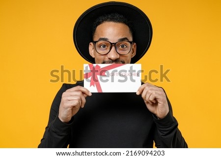 Young happy surprised fun african man 20s wear stylish black shirt hat tattoo translate fun eyeglasses cover mouth with gift voucher flyer mock up isolated on yellow orange background studio portrait.