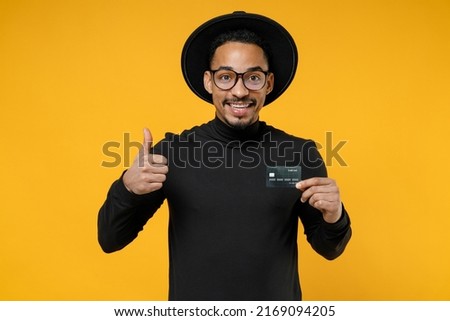 Young happy smiling successful american african man 20s in stylish black shirt hat eyeglasses holding credit bank card show thumb up like gesture isolated on yellow orange background studio portrait. Royalty-Free Stock Photo #2169094205