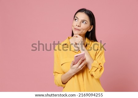 Young brunette pensive dreamful wistful latin happy cute woman 20s in yellow shirt hold mobile cell phone chat typing sms looking aside prop up chin isolated on pastel pink background studio portrait Royalty-Free Stock Photo #2169094255