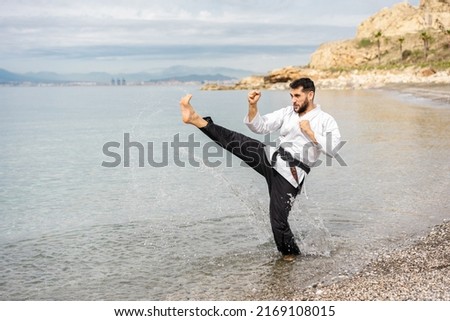 kung fu expert wearing a kimono and a black belt with the word "bushido" written in Japanese, performing a side kick on the shore of the beach splashing drops of water.  Royalty-Free Stock Photo #2169108015