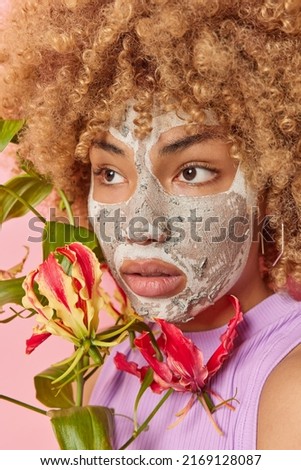 Close up shot of curly haired thoughtful woman looks somewhere applies beauty mask on face for skin treatment holds exotic plant dressed in casual clothes. Wellness spa and facial care concept