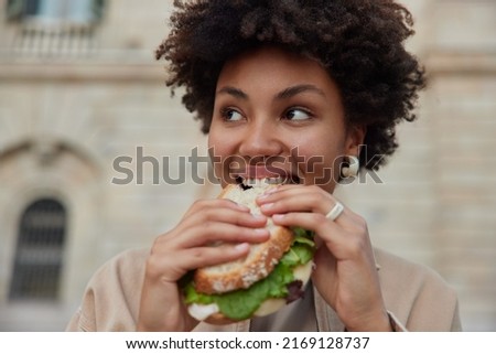 Pretty curly haired woman bites delicious sandwich poses outdoors at street looks away dressed casually has quick snack while walking outside being hungry. People lifestyle and fast food concept Royalty-Free Stock Photo #2169128737