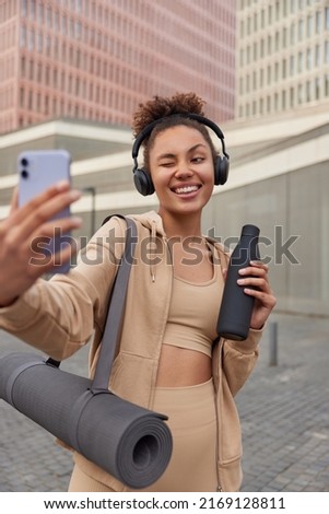 Vertical shot of happy sporty young woman takes selfie via smartphone winks eye holds bottle of water carries rolled fitness mat listens music via headphones stands against modern buildings.