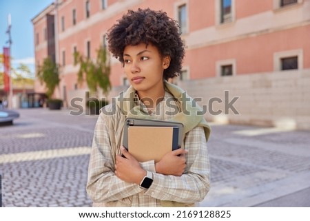 Thoughtful young woman carries paper notebook and portable tablet dressed in casual clothes looks somewhere poses outdoors in urban setting returns from university. People and lifestyle concept