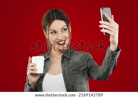 Young woman taking selfie on red studio background holding white plastic cup, smiling at phone camera, female blogger sharing content of everyday life with her followers on smartphone application