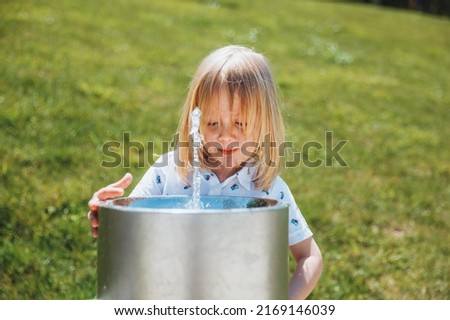 A little blond boy drinks from a drinking fountain in a city park on a hot summer day outdoors.