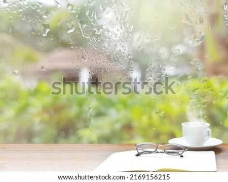 Rainy atmosphere with a drop of water on the glass. (a rainy day window background) 
On the table there is diaries, notebook glasses and white coffee mugs to save your holiday in Thailand.