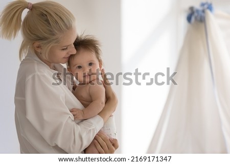 Young mother standing in cozy nursery cuddles, lulls her adorable baby in diaper, copy space. Happy motherhood and maternity, cherish, unconditional love, ad of goods and furniture for infants concept Royalty-Free Stock Photo #2169171347