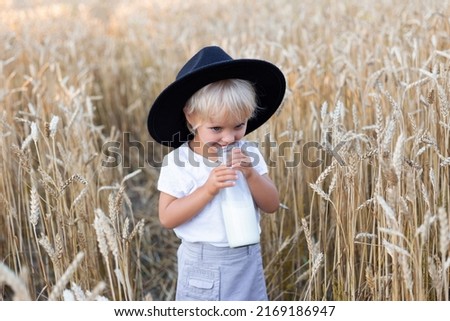 Happy cute cheerful caucasian little girl wearing hat and walking in summer wheat field holding a bottle of milk.
 Beautiful girl portrait outdoors. Childhood, beauty, happiness concept.
