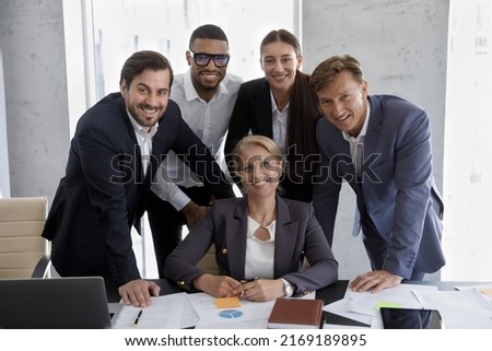 Staff in formal suits pose for corporate picture, gathered together in modern office board room. Middle-aged female boss, subordinates smile look at camera. Teamwork, business success, unity concept