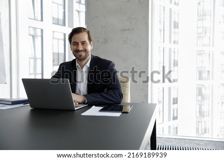 Portrait of successful businessman, business owner or manager in formal suit sit at desk with laptop smile looks at camera, feels satisfied by career growth in company. Business aspirations concept