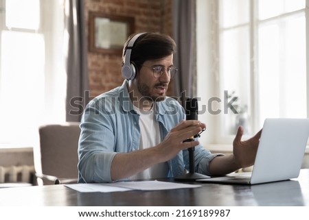Millennial man streamer wear headphones take part in virtual communication, records podcast seated at desk, speaking reflections on topic, make speech into microphone, create audio file. Tech concept