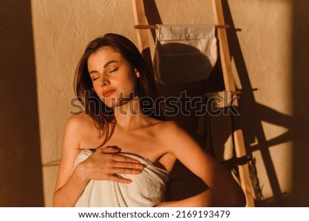 A young woman is enjoying the sun. A beautiful female model wrapped in a white towel stands in the bathroom after a shower. Shadows from tropical leaves. Royalty-Free Stock Photo #2169193479