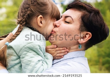 A mother with her eyes closed, kisses her little daughter on the cheek in the open air. A little daughter hugs her mother by the neck. Maternal care and love. Moments of tenderness.Happy loving family