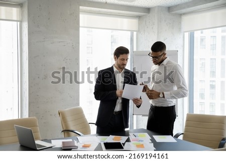 Two multi ethnic businessmen speaking in office, consider documents, share information, planning collaborative project, working together in modern board room. Teamwork, workflow and business concept Royalty-Free Stock Photo #2169194161