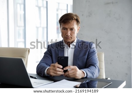 Successful serious middle-aged businessman using smartphone seated at workplace in skyscrapers office. Modern tech, business application usage, corporate messaging, agenda, appointments memo concept Royalty-Free Stock Photo #2169194189