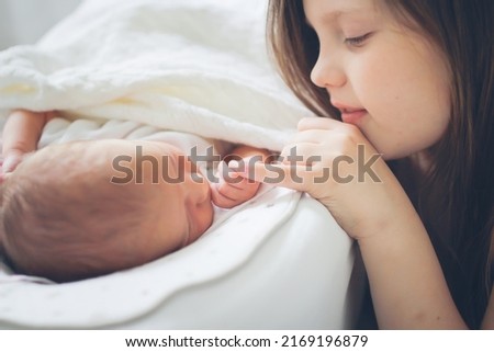 Cute European sibling children, newborn baby and older sister. Gentle touch and care. Girl child looks at a newborn baby at home in a bright bedroom.