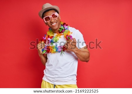 Handsome young man in Hawaiian necklace looking happy while standing against red background
