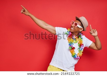 Happy young man in Hawaiian necklace pointing away while standing against red background