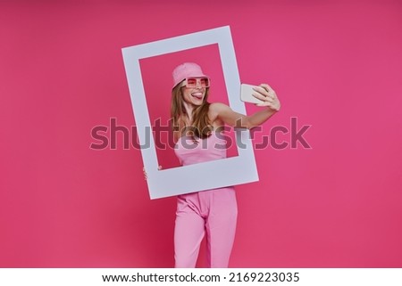 Playful woman making selfie and looking through a picture frame while standing against pink background