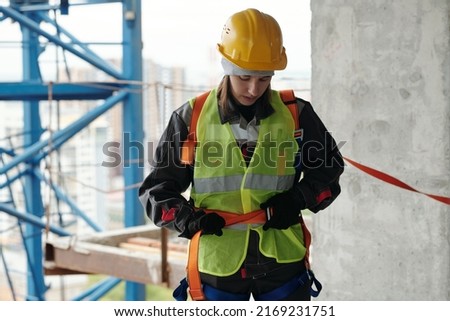 Young female builder in protective helmet and reflective vest fastening safety belt on waist before building work at construction site Royalty-Free Stock Photo #2169231751