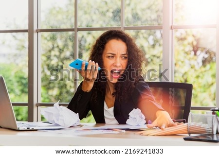 Businesswoman sitting at her busy  stress  tired desk shouting loudly while using her smartphone to talk to an overly rushed coworker irritates her when she delays deadline work. Royalty-Free Stock Photo #2169241833
