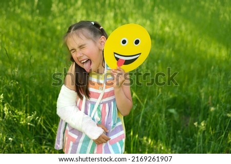 A bully child with a broken arm and a cast makes a grimace holding a smile face in his hand showing his tongue. A very emotional child. Emoji Day