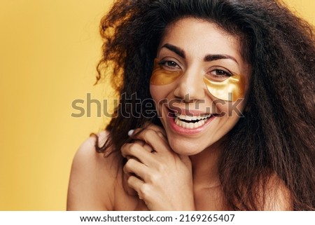 Happy curly pretty Latin female with gold hydrogel patches under eye laugh at camera posing isolated over yellow background. Cosmetic product ad Natural beauty concept Studio portrait