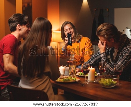 Laughing young woman showing funny selfie of her boyfriend to him and couple of friends during double date dinner at home
