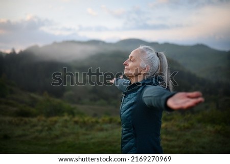 Senior woman doing breathing exercise in nature on early morning with fog and mountains in background. Royalty-Free Stock Photo #2169270969