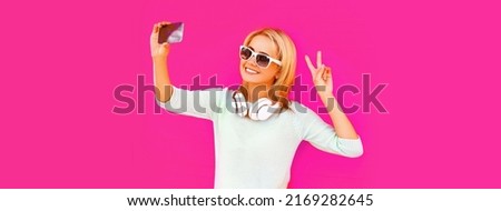 Portrait of happy smiling young woman taking selfie with smartphone in headphones listening to music on pink background