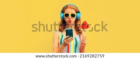 Summer colorful portrait of stylish young woman in headphones listening to music on smartphone with juicy lollipop or ice cream shaped slice of watermelon on blue background
