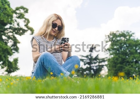 young Caucasian girl sitting on the grass and checking social media in the park. full shot. High quality photo